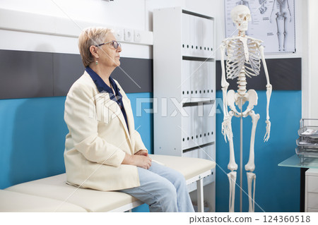 Older woman dressed casually sits in a hospital room prepared for her health consultation. Senior caucasian lady waits on examination bed for medical checkup in doctors office. 124360518
