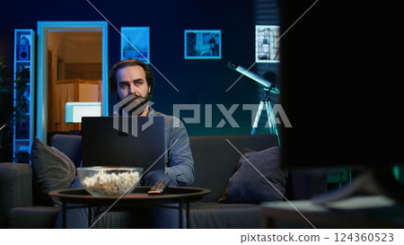 Man watching TV in apartment office and solving business related tasks on laptop, working from home. Freelancer on couch in living room looking at movies and working on notebook device, camera B Man watching TV in apartment office and solving business related tasks on laptop, working from home. Freelancer on couch in living room looking at movies and working on notebook device, camera B 124360523
