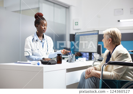 African american doctor consults with senior patient in wheelchair and reviews her medical history on computer. Female physician discusses treatment plans for white old woman during consultation. African american doctor consults with senior patient in wheelchair and reviews her medical history on computer. Female physician discusses treatment plans for white old woman during consultation. 124360527