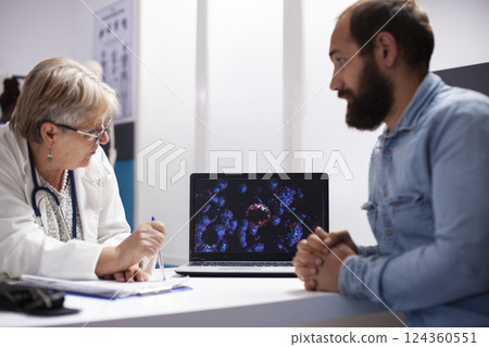 Old female physician and young male patient use laptop to analyze virus cells during consultation. Senior doctor with digital device illustrating enlarged microscopic details of germs to bearded man. Old female physician and young male patient use laptop to analyze virus cells during consultation. Senior doctor with digital device illustrating enlarged microscopic details of germs to bearded man. 124360551