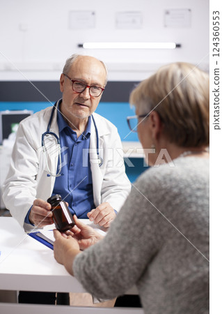 Older female patient sits with senior male physician in clinic, receiving prescribed bottle of pills for her recovery plan. Caucasian old doctor provides medicine to retired woman after medical exams. Older female patient sits with senior male physician in clinic, receiving prescribed bottle of pills for her recovery plan. Caucasian old doctor provides medicine to retired woman after medical exams. 124360553