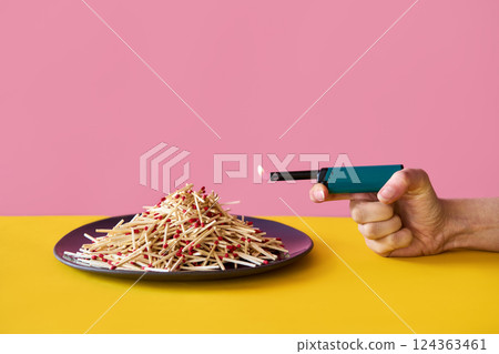 Hand with lit lighter pointed at pile of wooden matches on plate 124363461