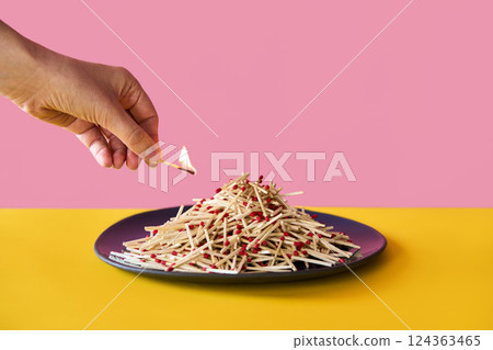 Hand holds burning matchstick above a large pile of matches on plate over vibrant background Hand holds burning matchstick above a large pile of matches on plate over vibrant background 124363465