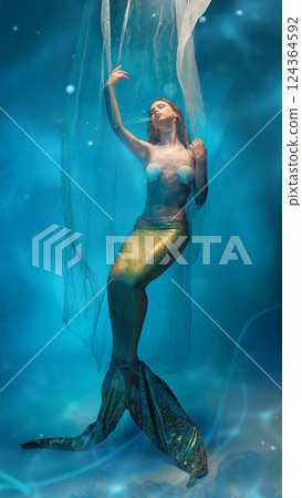 Graceful young woman, mermaid, caught between dreams and reality, extends hand toward light in surreal underwater world. 124364592