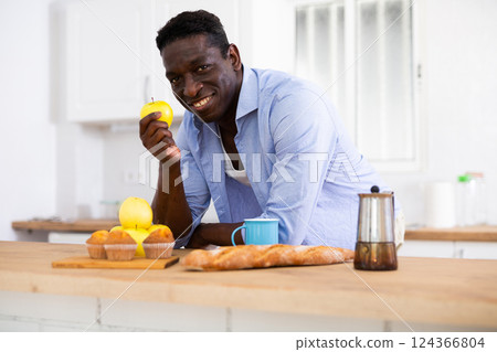 Smiling african american man eating apple in home kitchen Smiling african american man eating apple in home kitchen 124366804