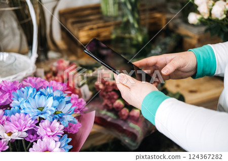 Florist using smartphone in flower shop to manage digital storefront. Reflects ecommerce floristry, small business tech, and mobile marketing trends Florist using smartphone in flower shop to manage digital storefront. Reflects ecommerce floristry, small business tech, and mobile marketing trends 124367282