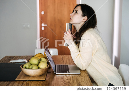 Frustrated woman holding notebook while staring upward during remote work struggle at kitchen table. Highlights work-from-home pressure and digital burnout 124367339