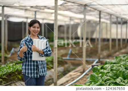 Confident female farmer stands in a greenhouse holding laptop and giving a thumbs up 124367650