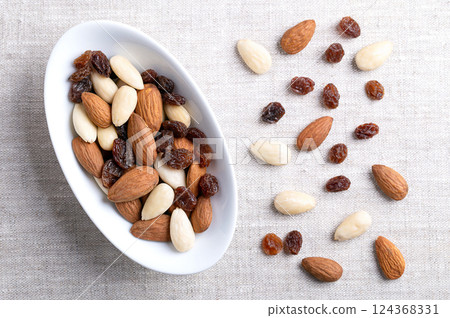 Almonds and raisins, a trail mix in an oval white bowl, on linen fabric. Also known as student food since 17th century, a mixture consisting of almonds with seed coats and blanched, and of raisins. Almonds and raisins, a trail mix in an oval white bowl, on linen fabric. Also known as student food since 17th century, a mixture consisting of almonds with seed coats and blanched, and of raisins. 124368331