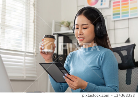 Design Review and Coffee Enjoyment. A woman in headphones examining design concepts while sipping coffee, illustrating a modern creative routine. 124368358