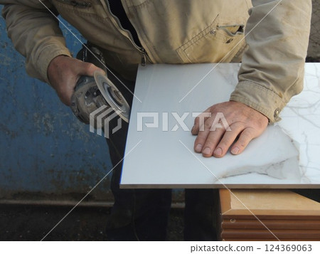 a craftsman with a grinder sharpens the edge of a large ceramic tile in an outdoor workshop, processing facing tiles with a round sharp disc on a grinder 124369063