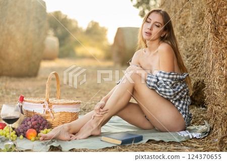 Young sexy woman in checked shirt sitting on rug with basket of fruit and wine on summer day among straw 124370655