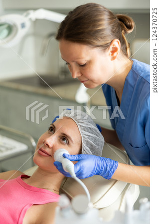 Female cosmetologist performing anti-aging face phototherapy procedure, shrink pores and repair damaged skin for young girl client in clinic of aesthetic medicine 124370725