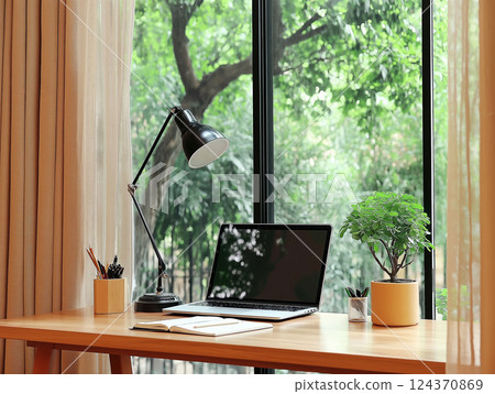 Minimalist home office setup with a laptop, desk lamp, and potted plant near a large window 124370869