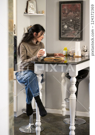 A woman is savoring a warm beverage as she immerses herself in the pages of a captivating book, surrounded by the cozy ambiance of her inviting kitchen. 124372369