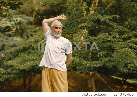 Senior man in white t-shirt standing at park and doing excercises. Positive mature woman has fitness or yoga class in nature. Wellness and healthy lifestyle on retirement. 124373466