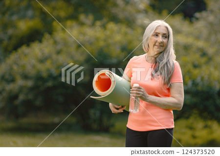 Senior woman with yoga mat standing at park with bottle of water. Positive mature woman after fitness or yoga class in nature. Wellness and healthy lifestyle on retirement. 124373520