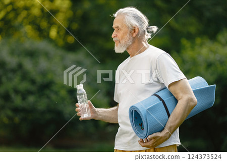 Senior man with yoga mat standing at park with bottle of water. Positive mature man after fitness or yoga class in nature. Wellness and healthy lifestyle on retirement. 124373524