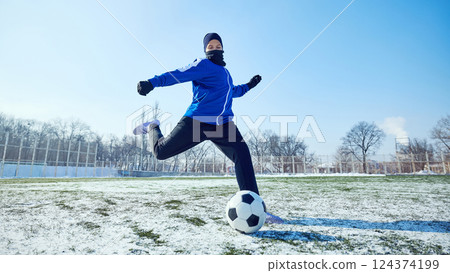 Dynamic Kick in Motion. Young football player in blue uniform powerfully kicking soccer ball on outdoor field covered with snow. 124374199