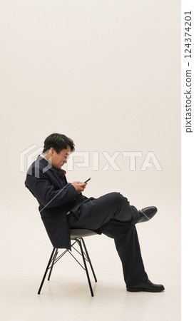 Side view photo of young Asian man sitting on chair and using smartphone, browsing or chatting against white background. 124374201