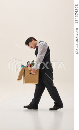 Young Asian man, dressed formal attire, leaving his job with heavy cardboard box, walking with sad expression against white background. Young Asian man, dressed formal attire, leaving his job with heavy cardboard box, walking with sad expression against white background. 124374208