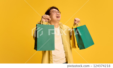 Portrait of happy, cheerful young Asian guy, dressed casual attire, and holding shopping bags with joyful expression against sunny yellow background. Portrait of happy, cheerful young Asian guy, dressed casual attire, and holding shopping bags with joyful expression against sunny yellow background. 124374218