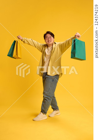 Happy Asian man walking, raising hands and showing many purchases after shopping against sunny yellow background. Happy Asian man walking, raising hands and showing many purchases after shopping against sunny yellow background. 124374219