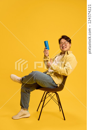 Happy young Asian man sitting on chair and showing thumb up and can with empty space against sunny yellow background. 124374221