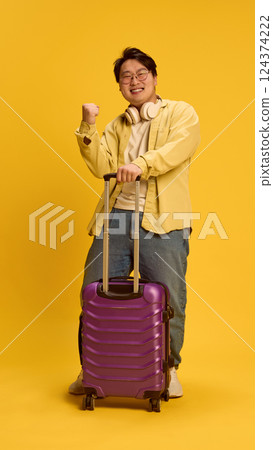Overjoyed young Asian man, traveler clenching fist of happiness he have nice holidays, standing next to huge bag against sunny yellow background. Overjoyed young Asian man, traveler clenching fist of happiness he have nice holidays, standing next to huge bag against sunny yellow background. 124374222