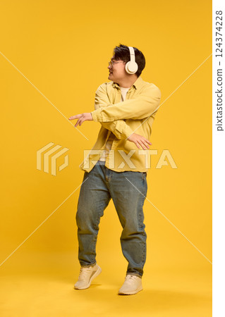Dynamic portrait of young Asian man dressed casual, dancing while listening music, exude joy and happiness against sunny yellow background. 124374228