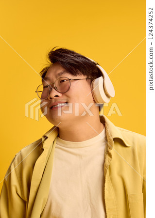 Close up photo of dreamy, overjoyed young Asian man, wearing glasses listening music in headphones against sunny yellow background. 124374252