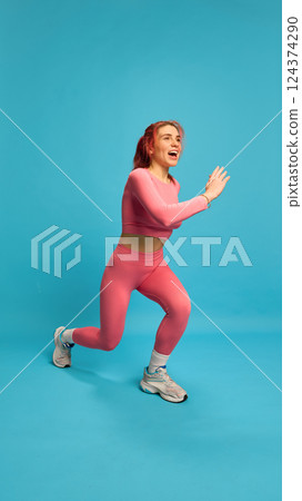 Smiling woman in pink activewear doing jumping lunge with powerful movement, embodying joy and effort in sport. 124374290
