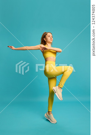 Energetic young woman in yellow activewear raising knee in balance pose, arms stretched, focusing on coordination and control. 124374805