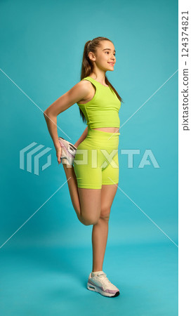 Full length photo of young girl, doing stretching exercises for legs, balancing on one leg against bright blue studio background. 124374821