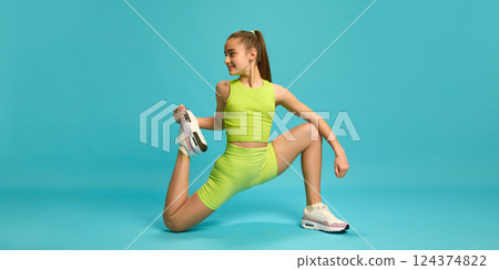 Flexible young woman doing exercises, stretching her legs, dressed bright green sportswear against blue studio background. Flexible young woman doing exercises, stretching her legs, dressed bright green sportswear against blue studio background. 124374822