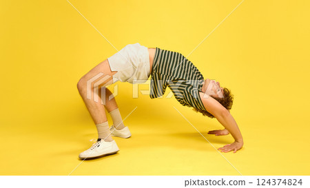 Young man dressed athleticwear, doing exercises for flexibility, bridge against bright yellow studio background. 124374824