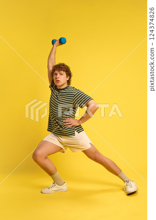 Confident and strong man, doing exercises for arms with dumbbells, dressed sporty outfit, against bright yellow background. 124374826