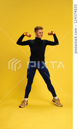 Fit man lifting dumbbells in biceps curl pose against yellow background, performing classic strength workout with confidence. 124374835