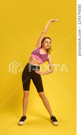 Young woman leaning sideways with one arm raised, engaging obliques in bright sporty outfit against bold yellow background. Young woman leaning sideways with one arm raised, engaging obliques in bright sporty outfit against bold yellow background. 124374880