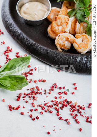Grilled big shrimps with parsley greens and sauce in a bowl on black cast iron plate on white surface and red pepper peas as a decoration. 124375156