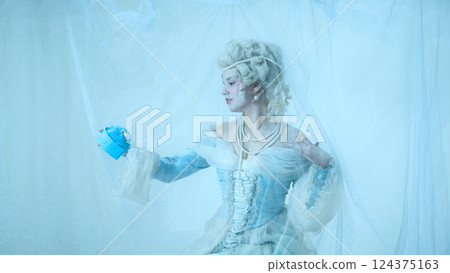 Portrait of graceful young woman as medieval duchess looking at luxurious necklace against light blue background. 124375163