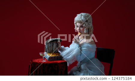 Tasting secrets. Portrait of baroque young woman in white dress eating chips from exotic chest, glamorous and surreal atmosphere Tasting secrets. Portrait of baroque young woman in white dress eating chips from exotic chest, glamorous and surreal atmosphere 124375184