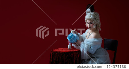 Playful ritual. Rococo-styled young woman pouring milk from blue jug into glass against deep red background. 124375188