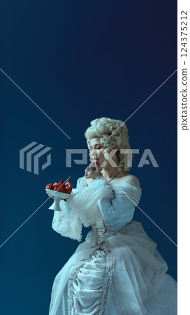 Regal elegance. Woman in white rococo gown with powdered wig enjoying pomegranate, stylized as royal figure on dark blue background. 124375212