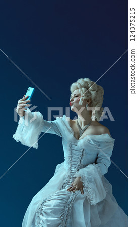 Selfie pose. Portrait of young woman in rococo look holding phone, imitating selfie trend on blue background. Selfie pose. Portrait of young woman in rococo look holding phone, imitating selfie trend on blue background. 124375215