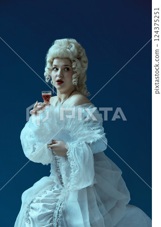 Surprised moment. Elegant woman holding glass with alcohol and fan, eyes wide in dramatic expression. 124375251