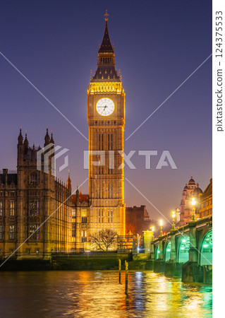 Big Ben stands tall against the night sky, illuminated in warm golden lights. The river reflects its beauty as the historic buildings create a stunning backdrop in London, England. Big Ben stands tall against the night sky, illuminated in warm golden lights. The river reflects its beauty as the historic buildings create a stunning backdrop in London, England. 124375533