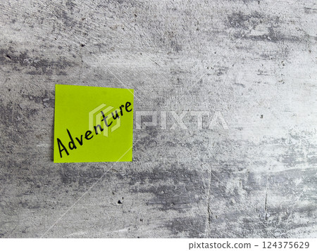 Vibrant yellow sticky note with word Adventure written on it, displayed against textured grey background, symbolizing creativity and inspiration for new experiences 124375629