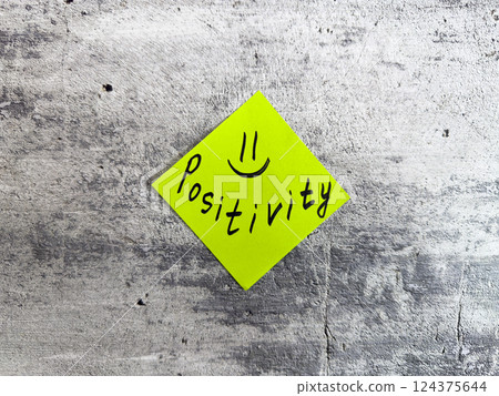 Yellow sticky note in a diamond shape displaying the word Positivity accompanied by a smiley face on a textured grey background, representing the essence of optimistic thinking and cheerful outlook 124375644