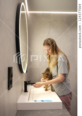 Teen girl preparing to bathe her reluctant cat at the sink in a modern bathroom with soft lighting and a round illuminated mirror. Teen girl preparing to bathe her reluctant cat at the sink in a modern bathroom with soft lighting and a round illuminated mirror. 124377004